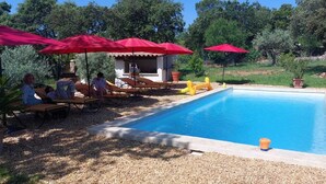 A swimming pool with lounge chairs and red umbrellas, surrounded by trees and a small building. - Location à la Semaine (Néoules)