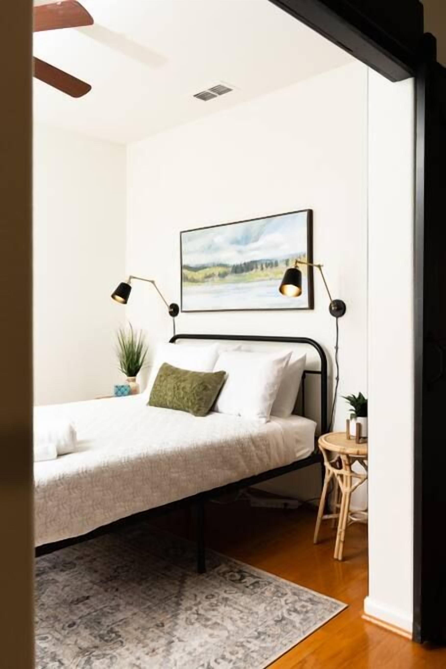 Une chambre à coucher avec un lit, une table de chevet, une lampe, un tableau accroché au mur et une petite plante en pot.