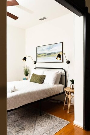 Quarto com cama, mesa de cabeceira, abajur, pintura na parede e uma pequena planta em vaso.