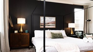 Een slaapkamer met een bed, bedlampjes, een nachtkastje, een lijst aan de muur en een raam met gordijnen.