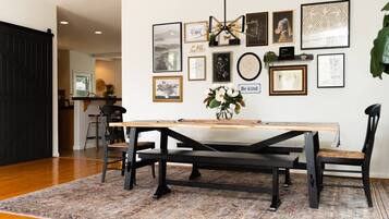 A dining area with a long table, black chairs, a patterned rug, and various framed artworks on the wall.