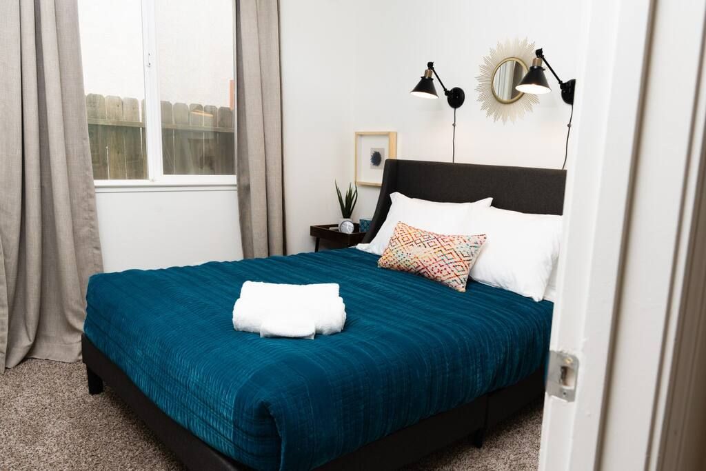 Un lit bien fait, recouvert d’une couvre-lit turquoise et agrémenté d’oreillers blancs, une petite table de chevet avec une lampe, et une fenêtre avec des rideaux.