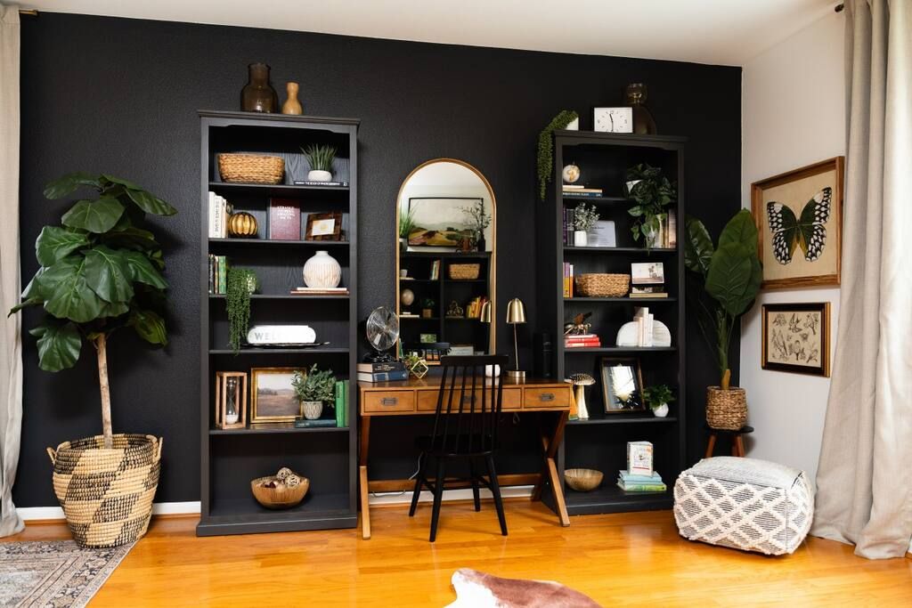 Een kamer met een bureau, twee boekenkasten, een spiegel en een grote plant.
