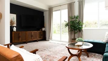 Un salon comprenant un canapé, une table basse, un téléviseur posé sur un meuble et une grande fenêtre avec des rideaux.