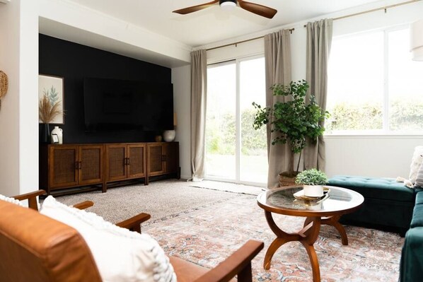 Una sala de estar con un sofá, una mesa de centro, un televisor sobre un mueble bajo y una ventana grande con cortinas.
