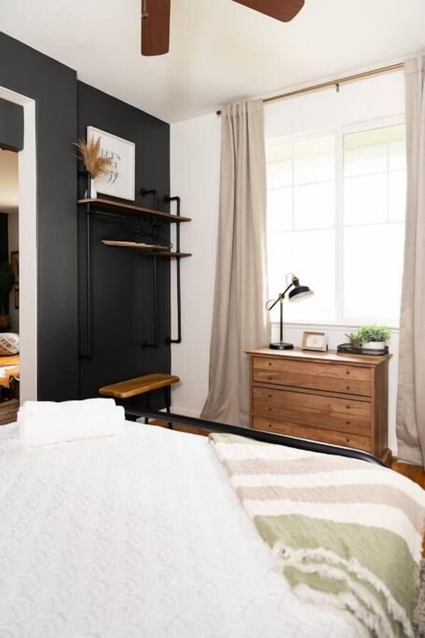 Une chambre à coucher comprenant un lit, une table de chevet, une commode, une fenêtre avec des rideaux et une étagère murale agrémentée d’une plante.