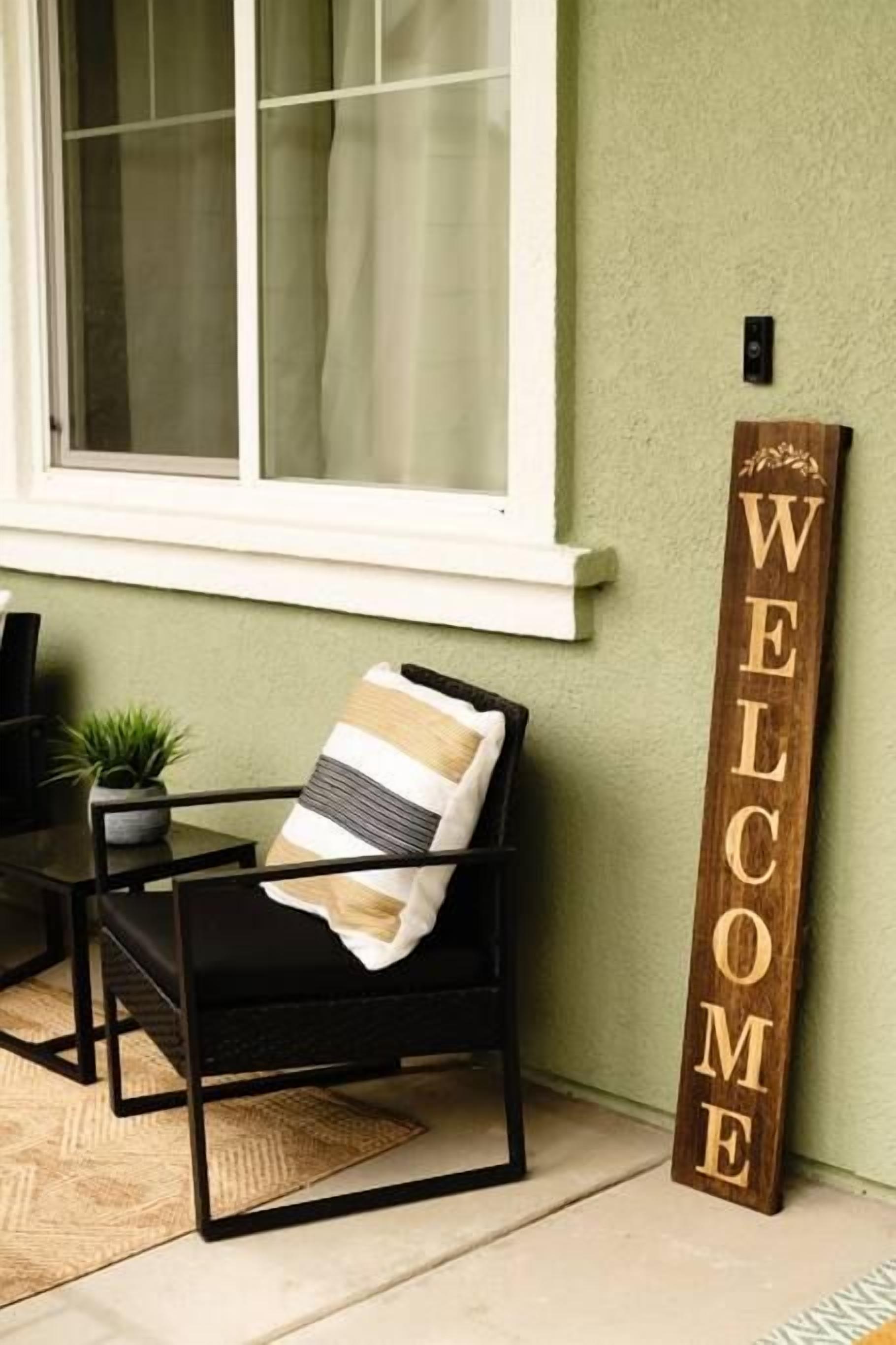Una silla de metal negro con un cojín a rayas, un letrero de madera con la palabra 'WELCOME' grabada, y una pequeña planta en maceta sobre una mesa.