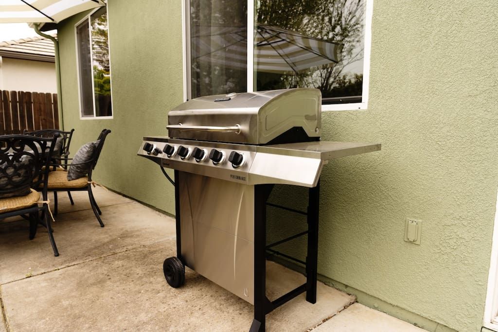 Un barbecue au gaz portatif, doté d’un panneau de commande, est placé sur une terrasse en béton adjacente à une maison dont le mur extérieur est peint en vert et qui possède une fenêtre.