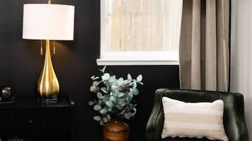 Um quarto com parede escura, poltrona verde, luminária, vaso com plantas e janela com cortinas.