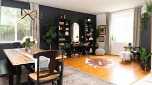 Een moderne woonkamer met een houten eettafel, donkere stoelen, een grote spiegel en een zwarte boekenkast vol decoratieve objecten.