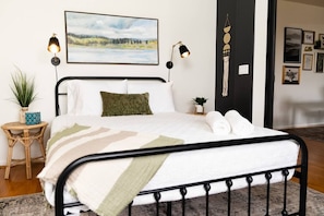 Una cama bien hecha con edredón blanco y manta verde, una mesita de noche con una planta y una lámpara de pared.