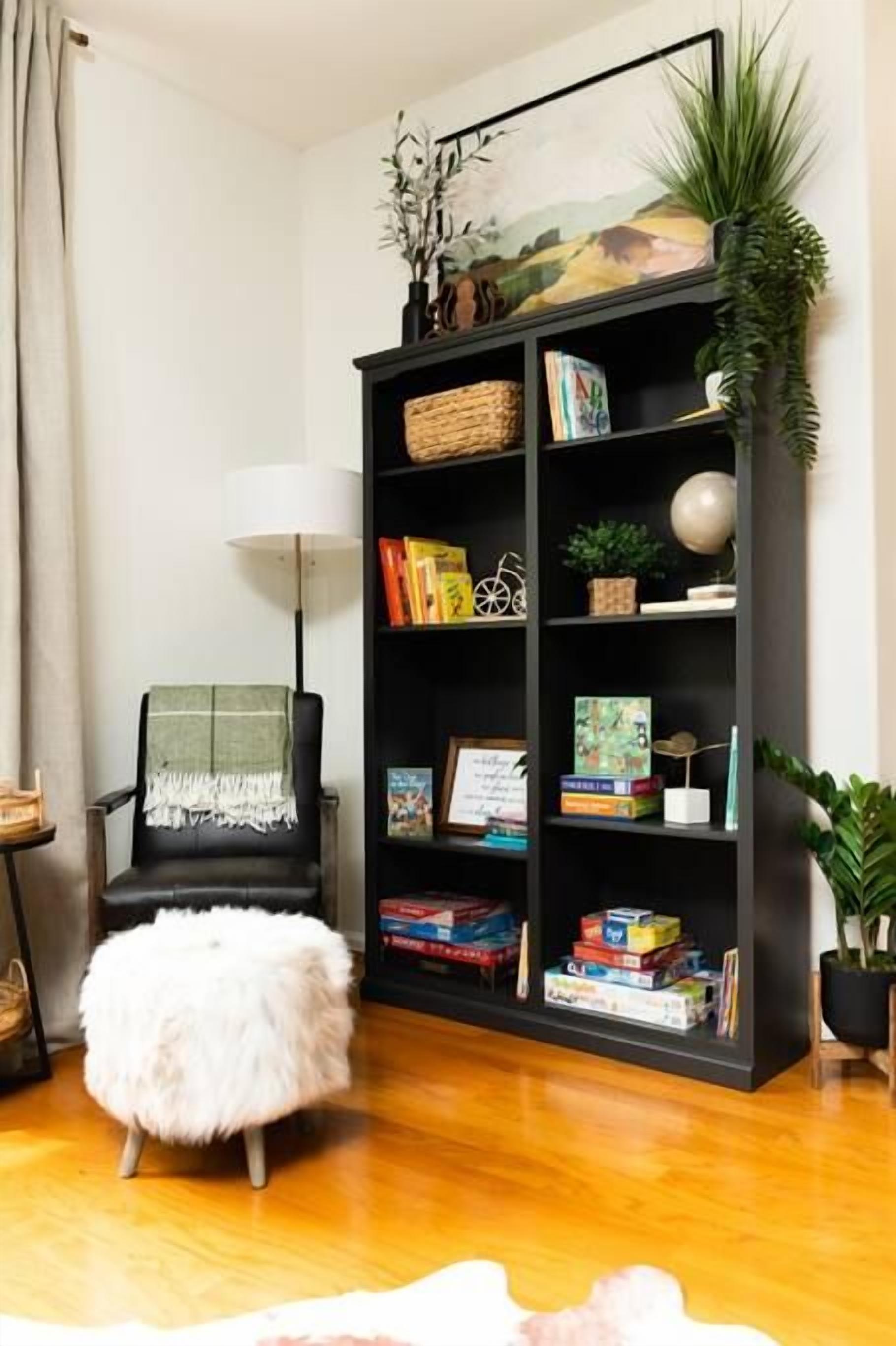 Una habitación con una estantería negra llena de libros y juegos, un otoman blanco y mullido, un sillón negro con una manta verde y una lámpara.
