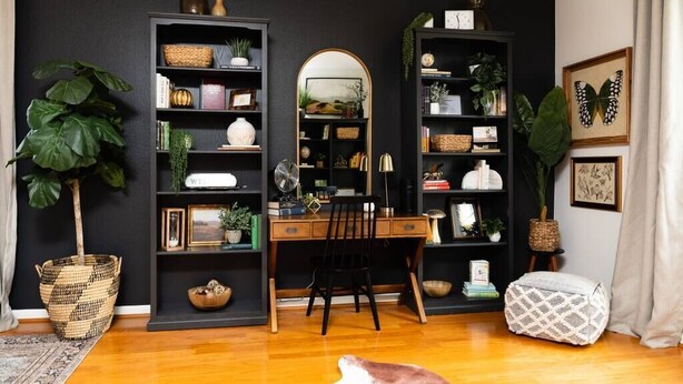 Een kamer met een houten bureau, twee zwarte boekenkasten, een spiegel en een grote plant.