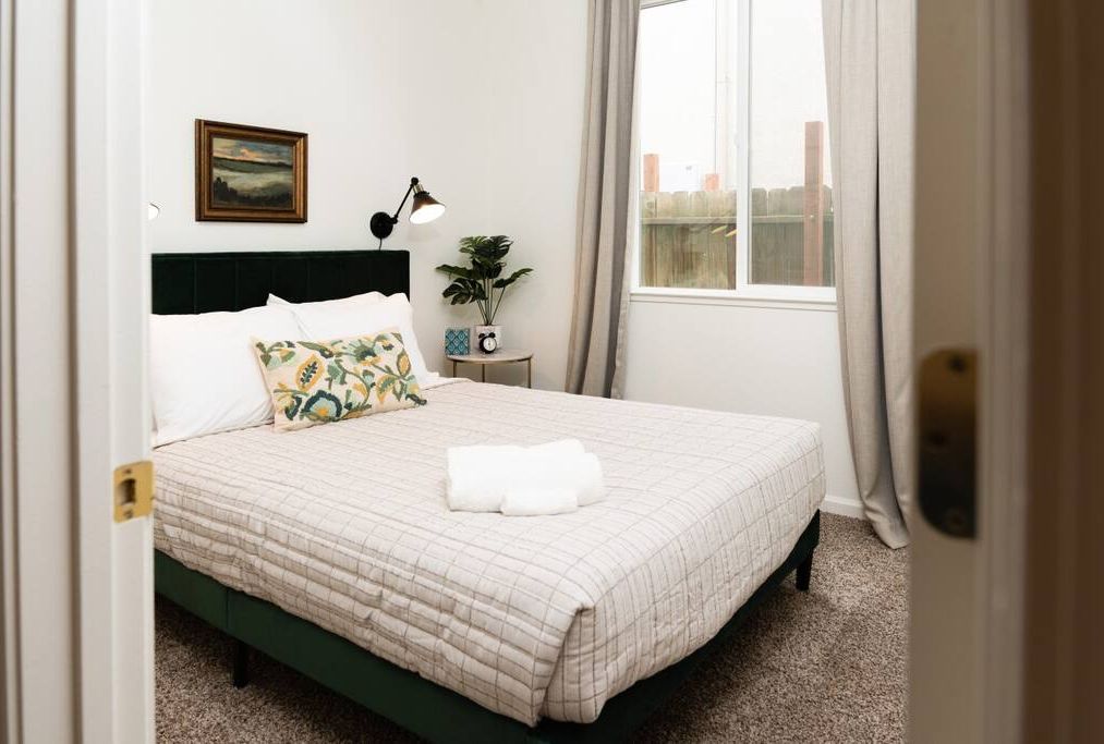 Una cama bien hecha con colcha acolchada, una lámpara de noche, una planta pequeña en maceta y un cuadro en la pared.