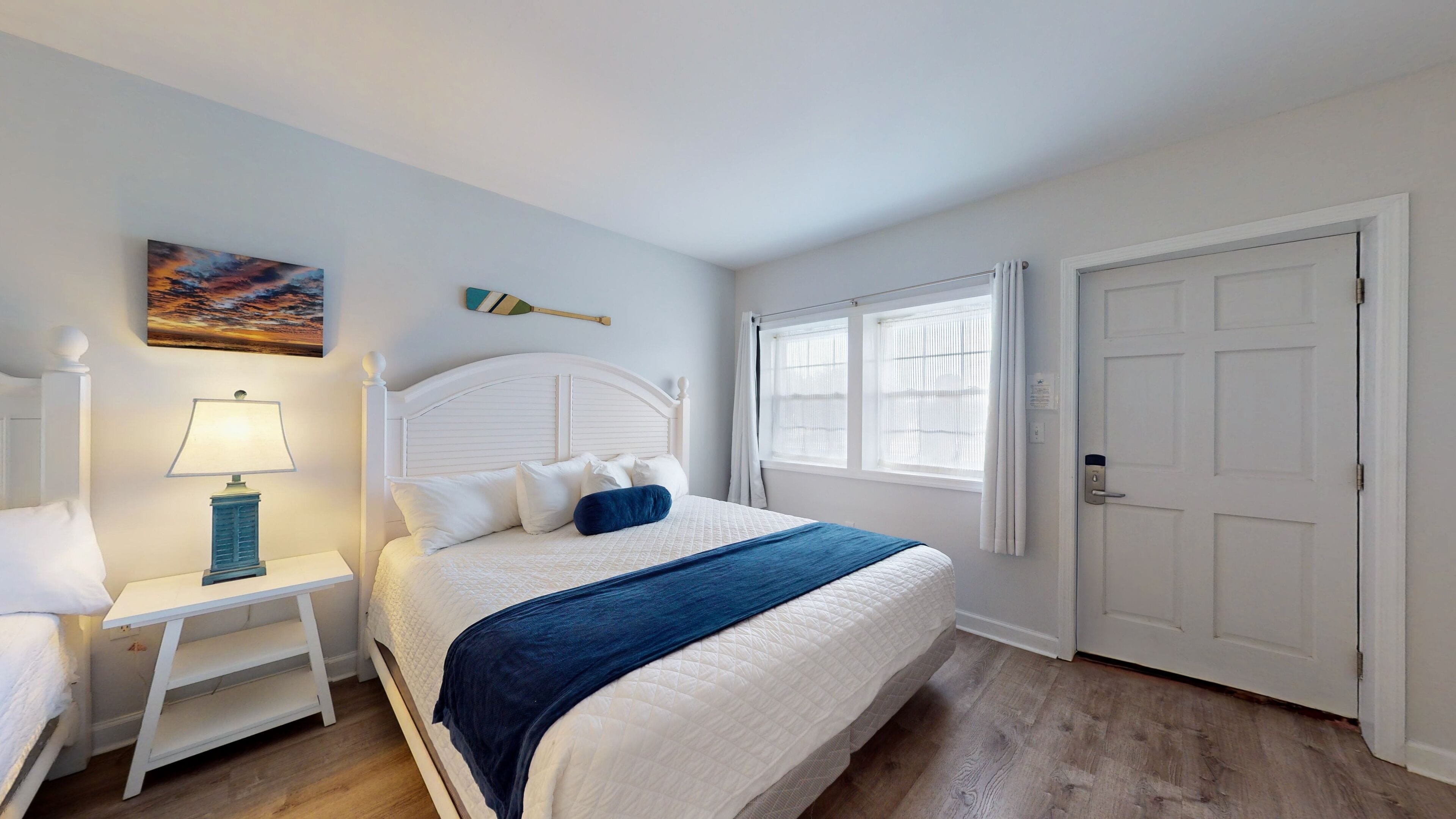 Une chambre à coucher avec un lit blanc, une table de chevet avec une lampe, un tableau au mur et une fenêtre avec des rideaux.