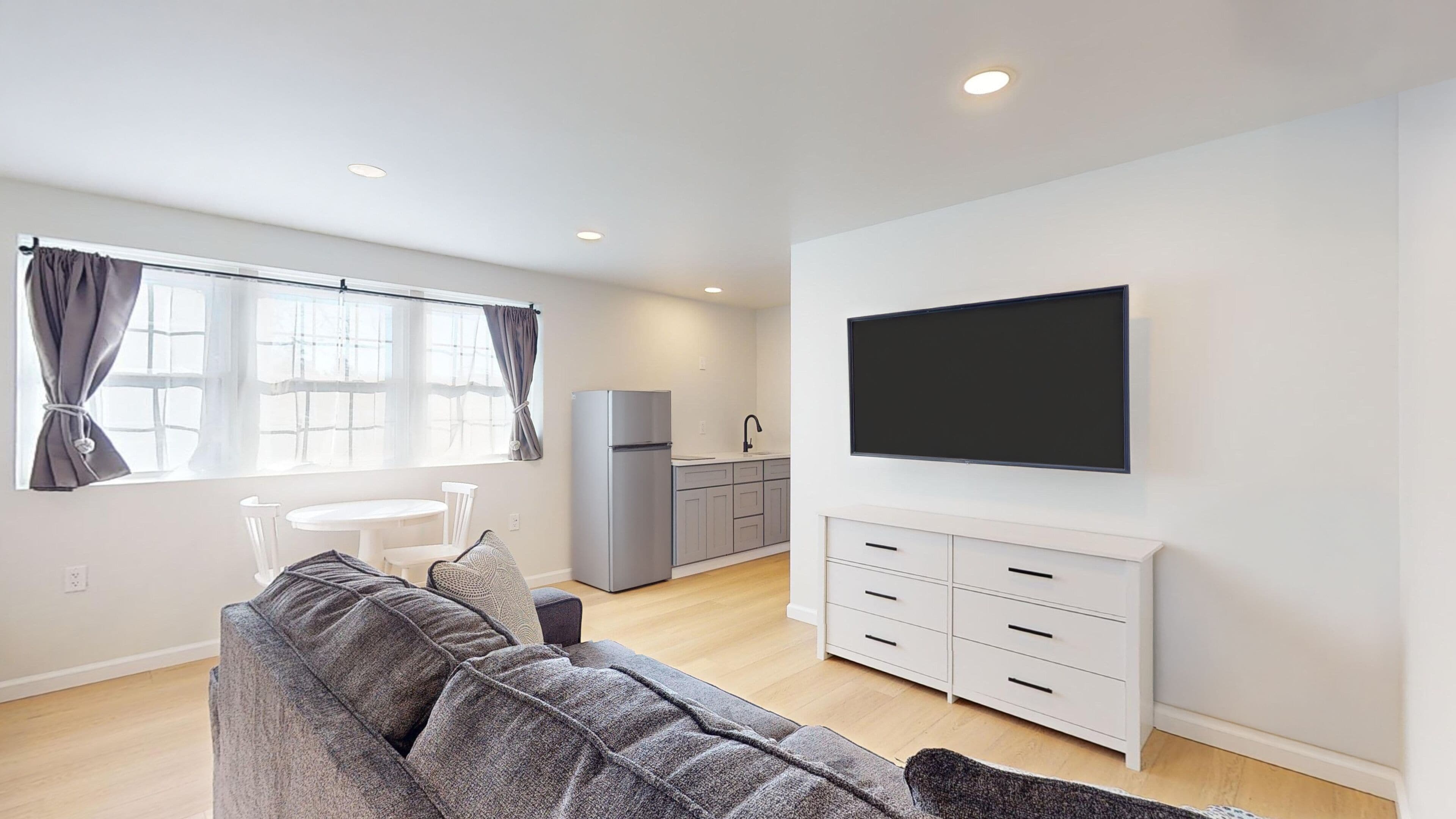 Una sala moderna con un sofá gris, un televisor de pantalla plana y una cocina con armarios grises y un refrigerador.