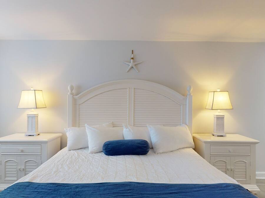 Un dormitorio con una cama grande, cabecera blanca, dos mesitas de noche con lámparas y una decoración de estrellas en la pared.