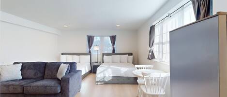 Una habitación de hotel moderna con un sofá gris, una cama con ropa blanca, una pequeña mesa con sillas blancas y un gran ventanal con cortinas.