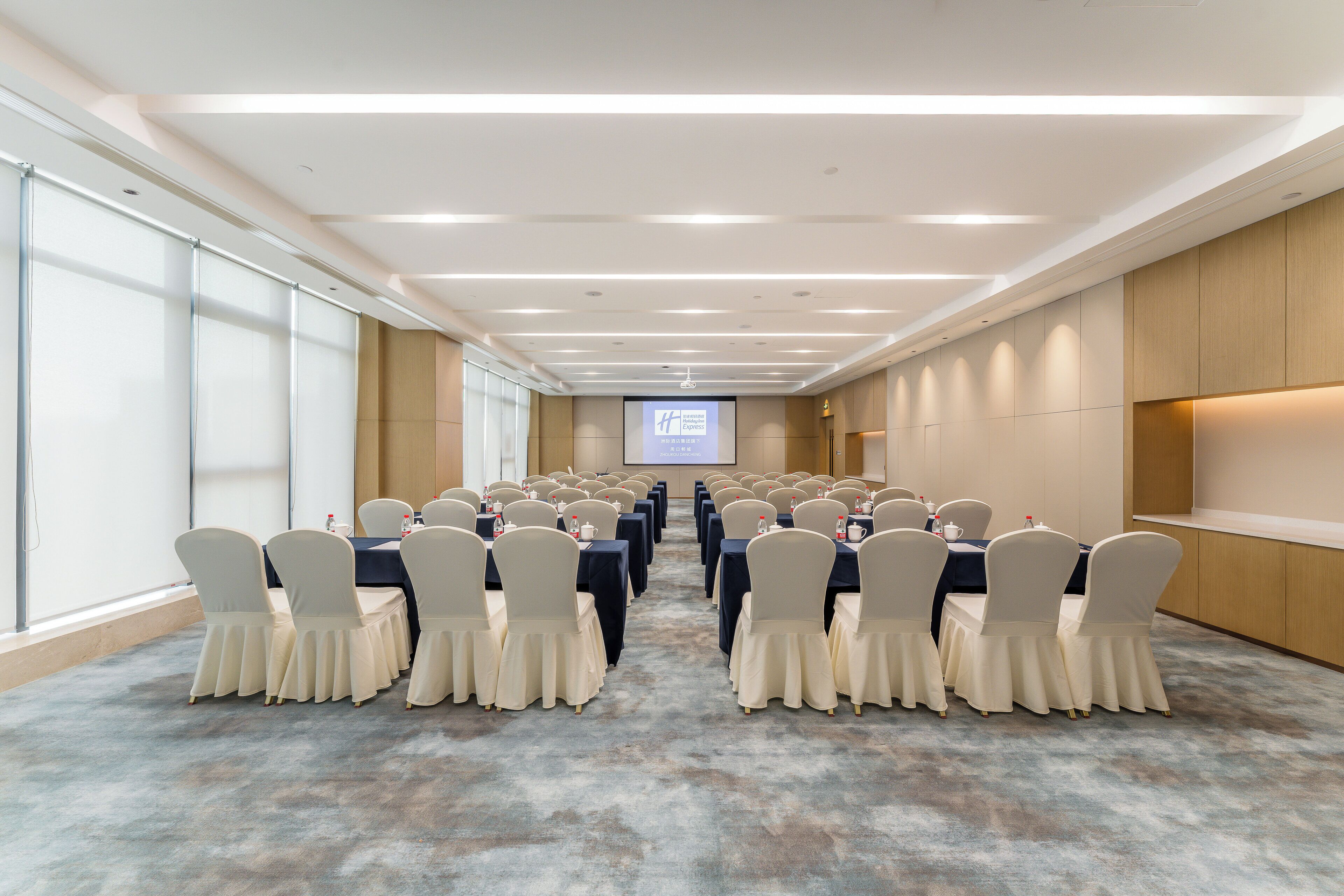 A conference room with rows of tables and chairs, a projector screen, and a carpeted floor.