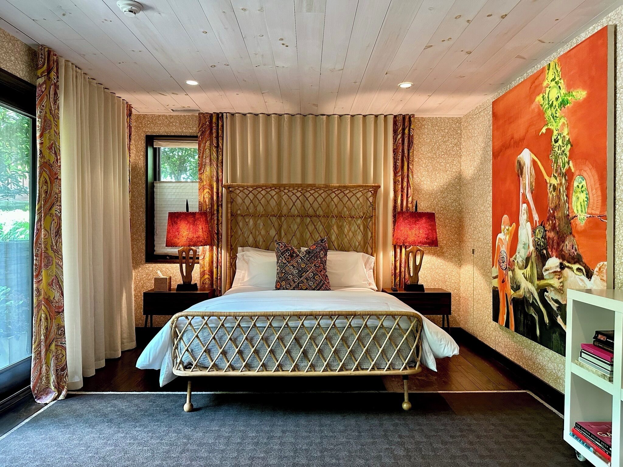 Quarto de hotel com uma cama grande, duas luminárias de cabeceira, uma janela com cortinas e um quadro grande na parede.