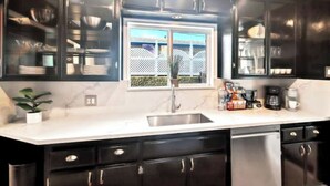 A modern kitchen with dark cabinets, a white countertop, and a window with a view of a white fence and greenery. - 7mins LAX/Beach - Gorgeous Home (Hawthorne)