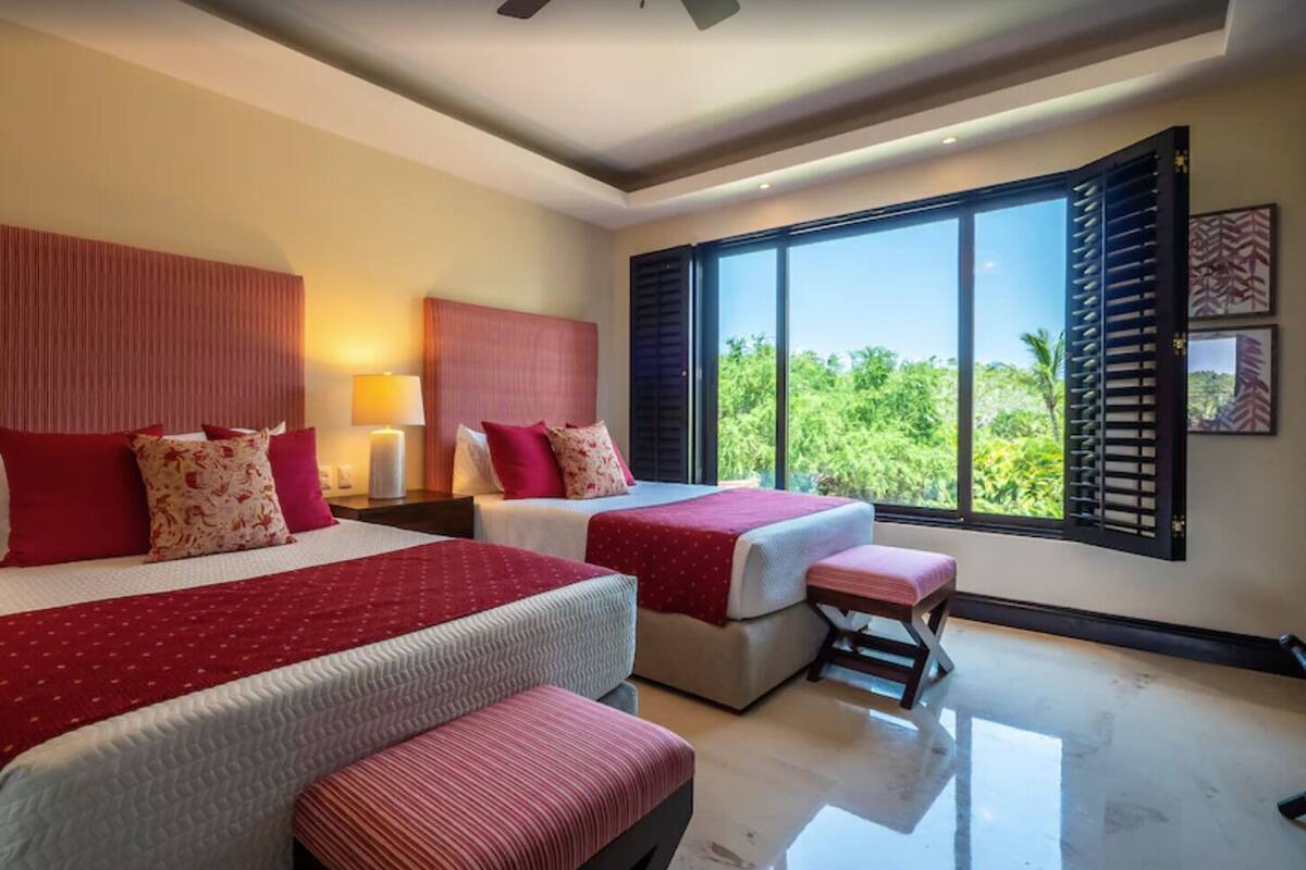 A modern hotel room with two beds, large windows, and a view of greenery.