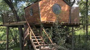 Ein Vogelhaus aus Holz mit einer Leiter und einem runden Fenster, umgeben von Bäumen.