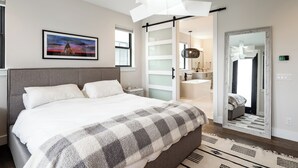 A modern bedroom with a large bed, a checkered throw blanket, a painting on the wall, and a bathroom visible through an open door. - 5-minute walk to downtown!   27-day minimum rental.  (Bozeman)