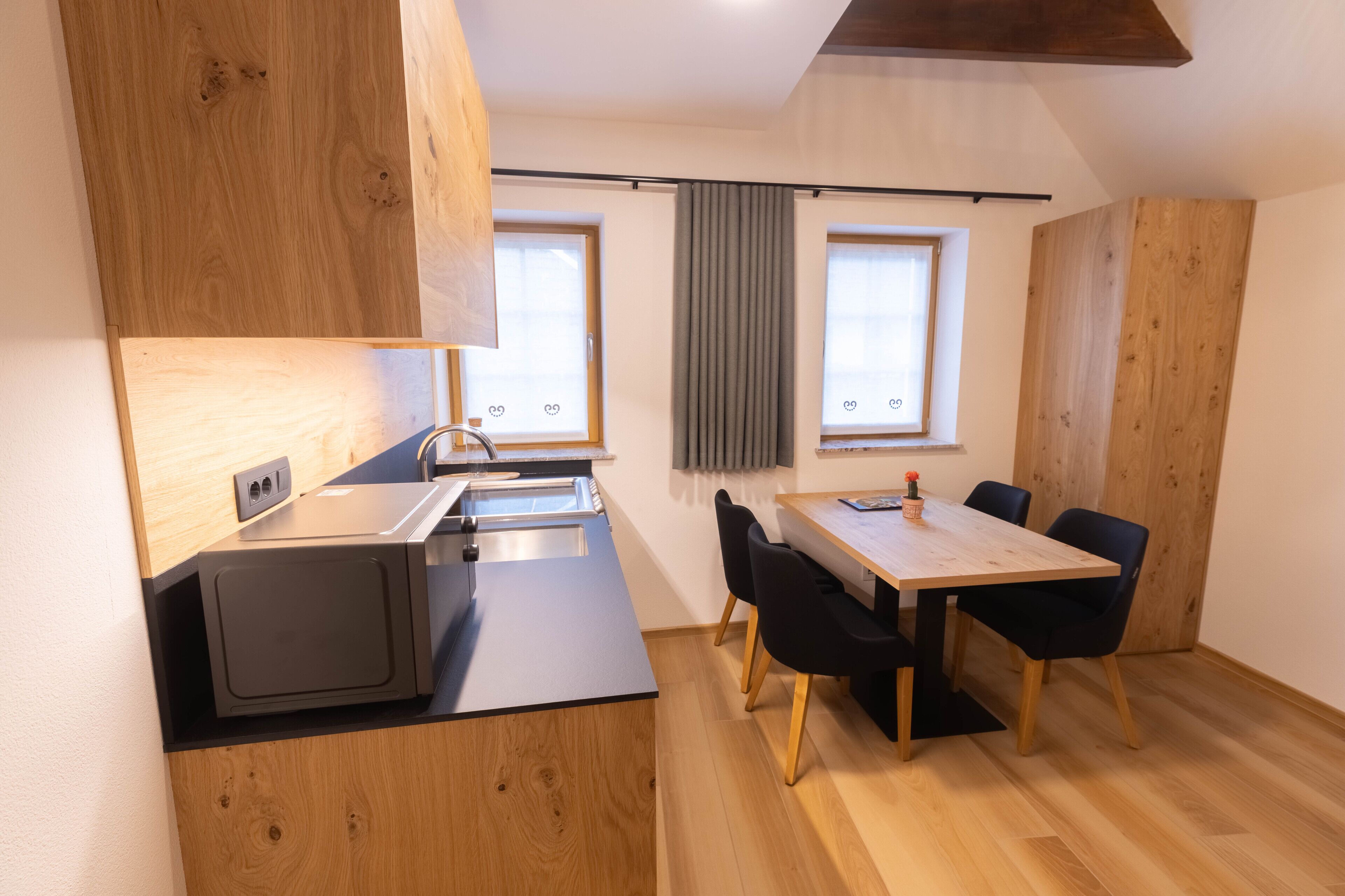A compact kitchen with a microwave, sink, and wooden cabinets. There is a dining table with chairs and a window with curtains.