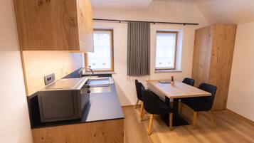 A compact kitchen with a microwave, sink, and wooden cabinets. There is a dining table with chairs and a window with curtains.