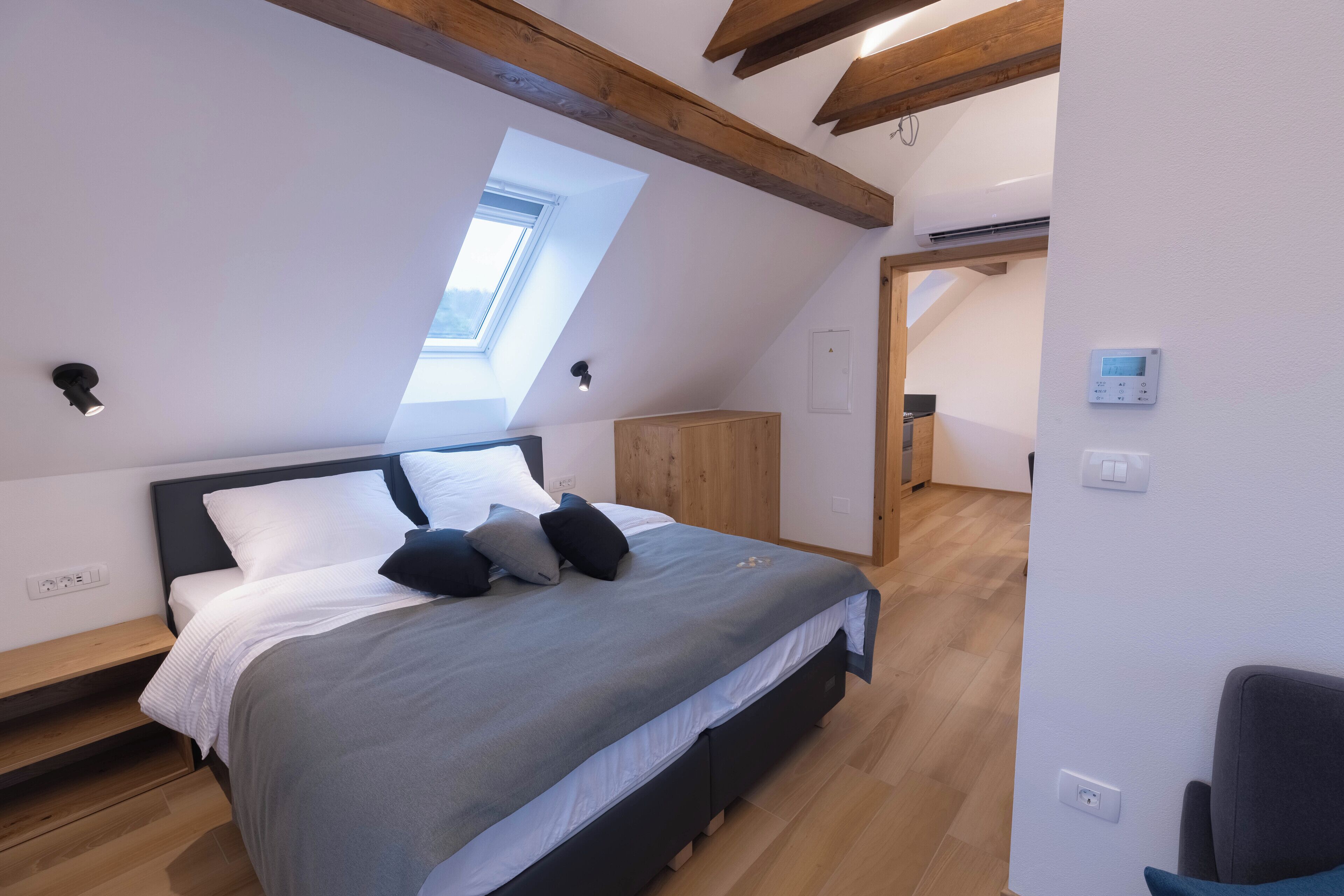 Een slaapkamer met een schuine kap, dakraam, bed met grijze plaid, houten hoofdbord en een klein houten kastje.