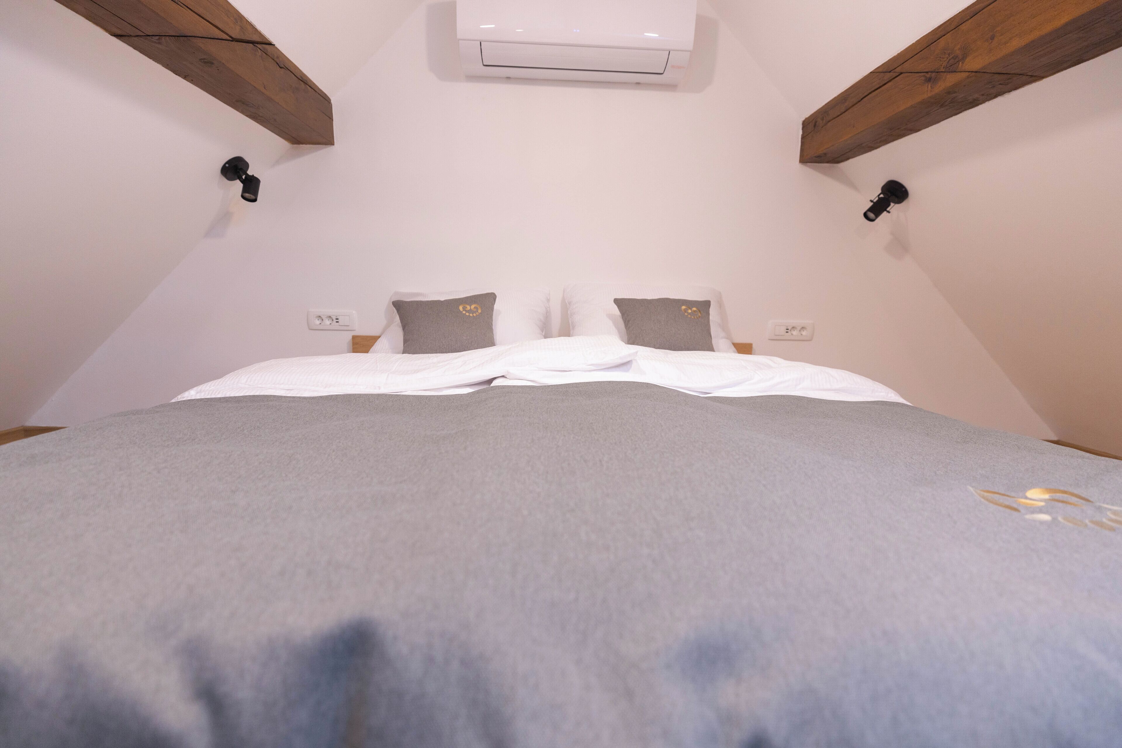 Una camera da letto con un letto, biancheria grigia, un condizionatore d'aria bianco, travi in legno e due interruttori a parete.