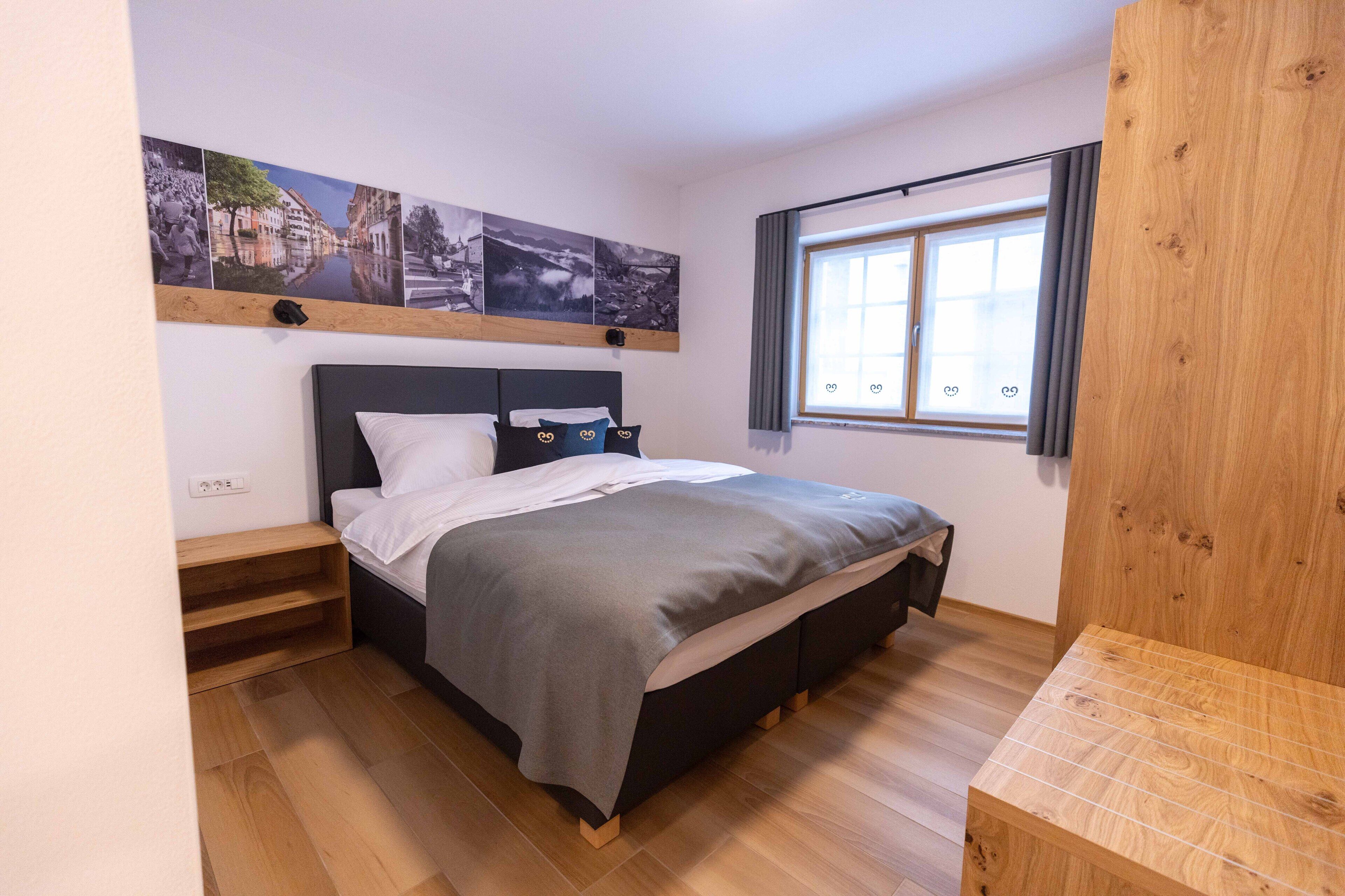 A bedroom with a bed, wooden floor, a window with curtains, and a wall-mounted shelf with pictures.