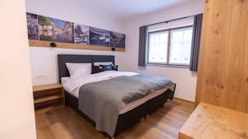 A bedroom with a bed, wooden floor, a window with curtains, and a wall-mounted shelf with pictures.
