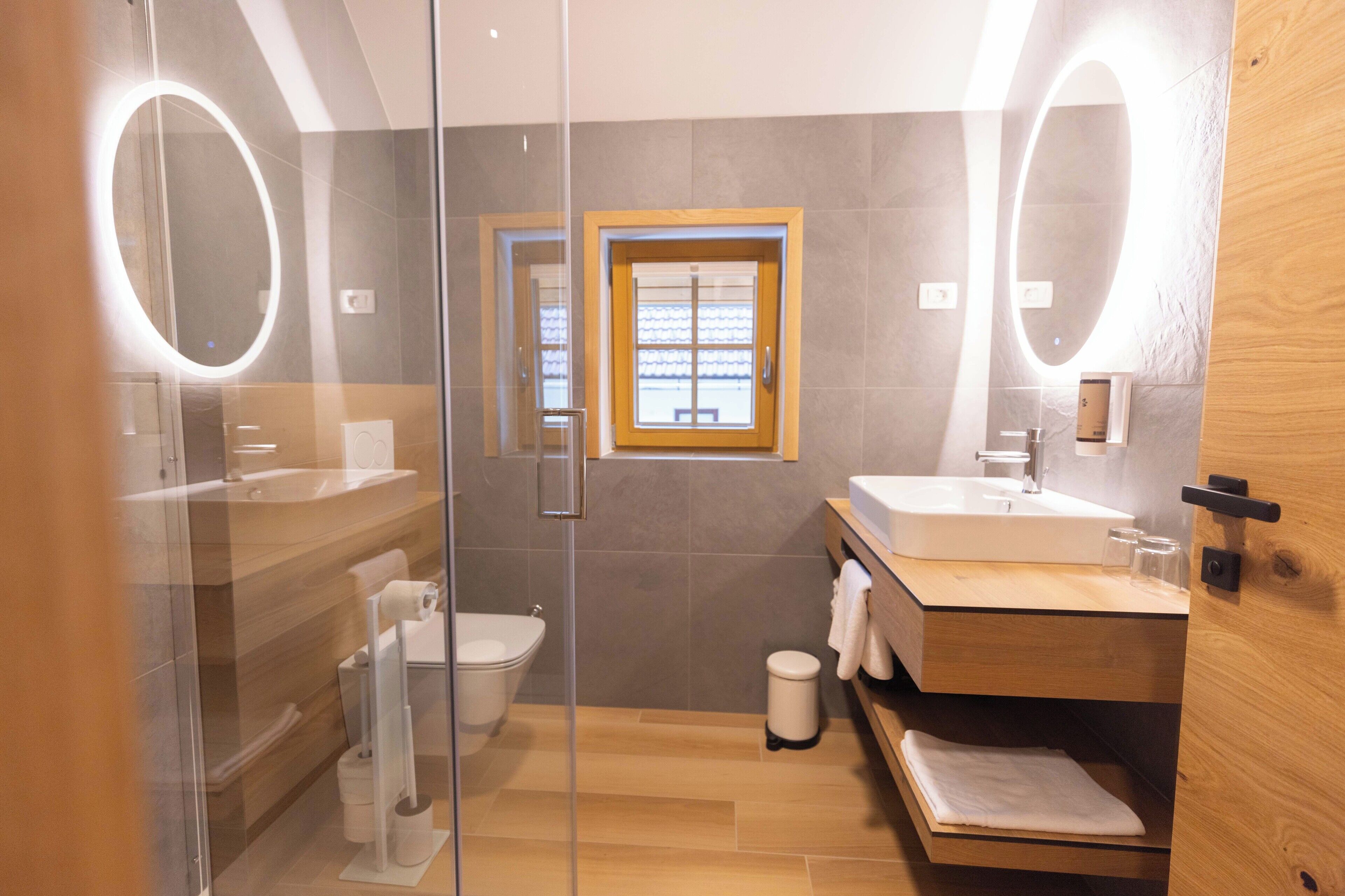 Un baño moderno con cabina de ducha de vidrio, tocador de madera con lavamanos y espejo con luz.
