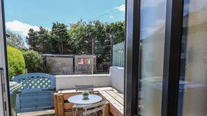 Pequeno pátio com mesa de madeira e cadeira dobrável, planta em vaso e vista para área cercada com árvores e um edifício ao fundo.