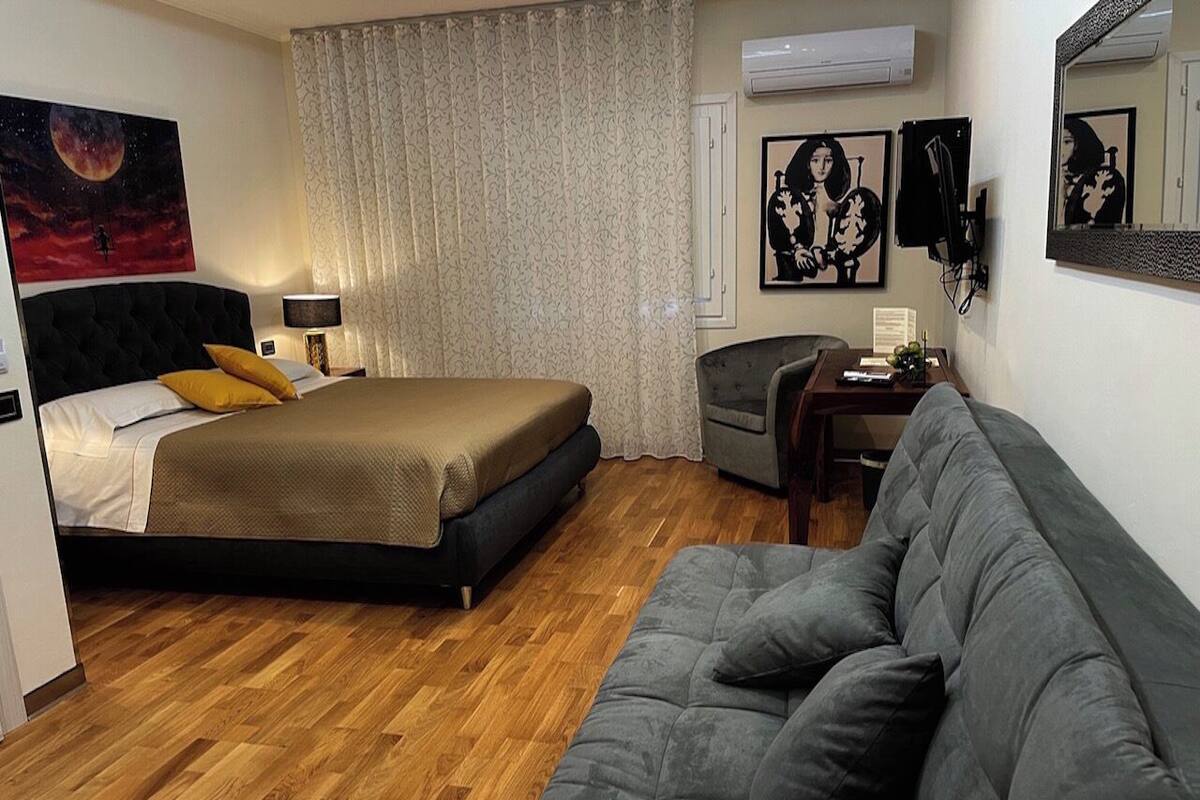 A hotel room with a bed, a grey sofa, a small desk, and a window with curtains.