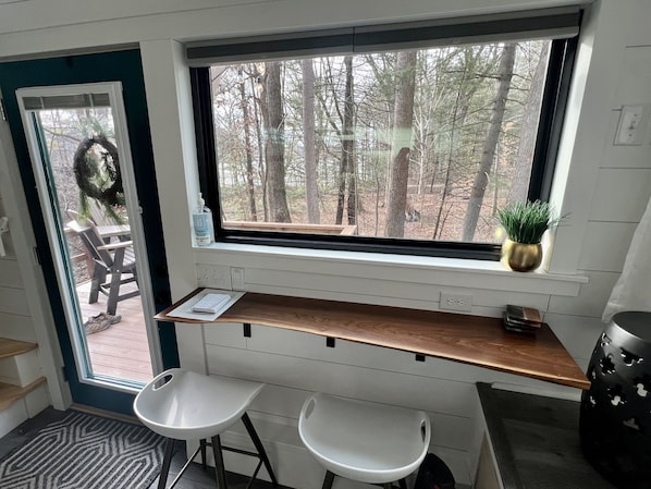 Una habitación con un amplio ventanal con vistas a un bosque, un escritorio de madera, dos taburetes blancos y una pequeña planta en maceta en el alféizar.