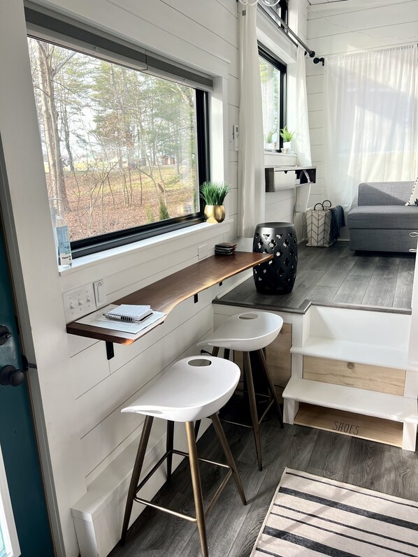 A modern interior with a window overlooking a forest, a small table with a notebook, two white bar stools, and a grey sofa.