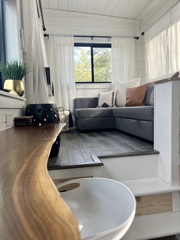 A modern living room with a wooden dining table, grey sofa, and a large window with curtains.