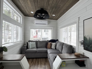 Ein modernes Wohnzimmer mit einem Ecksofa, Holzboden, großen Fenstern mit Jalousien, einem Deckenventilator und einem kleinen Tisch mit einer Pflanze.