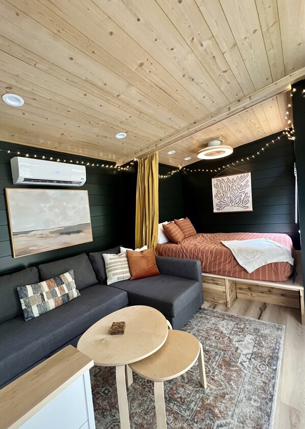 A cozy room with a wooden ceiling, a bed with a striped blanket, a gray sofa with multiple cushions, a small round table, and a patterned rug.
