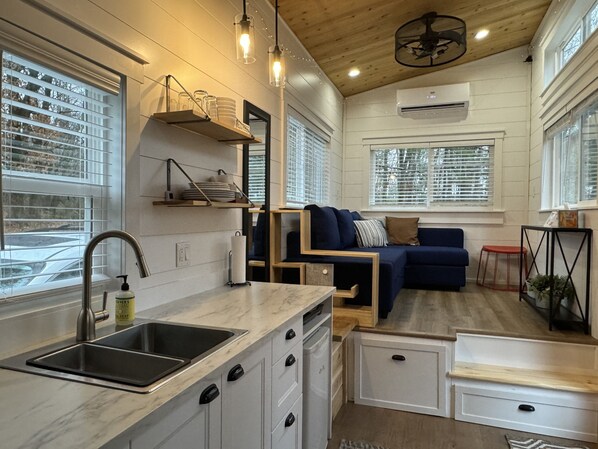 Cocina pequeña con lavabo, alacenas de almacenamiento y encimera. Hay un sofá azul y una mesita con una planta.
