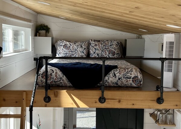 A loft bedroom with a bed, a ceiling fan, a window, and a plant.