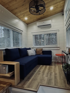 A modern living room with a dark blue sectional sofa, wooden flooring, and a ceiling fan. - The Gladiola Tiny House by the Lake (Logan)