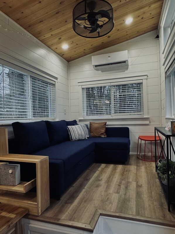 A modern living room with a dark blue sectional sofa, wooden flooring, and a ceiling fan. - The Gladiola Tiny House by the Lake (Logan)