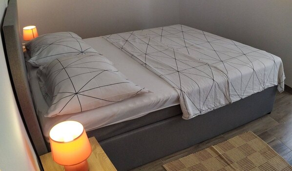 Una cama bien hecha con una colcha y almohadas de estampado geométrico, una lámpara de noche y una alfombra tejida sobre un suelo de madera.