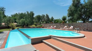 Ein rechteckiger Swimmingpool mit einer Steinmauer und einer Pflasterterrasse. Liegestühle mit roten Kissen laden zum Entspannen ein, und ein kleiner Gartenbereich mit Pflanzen und Blumen sorgt für eine grüne Note.