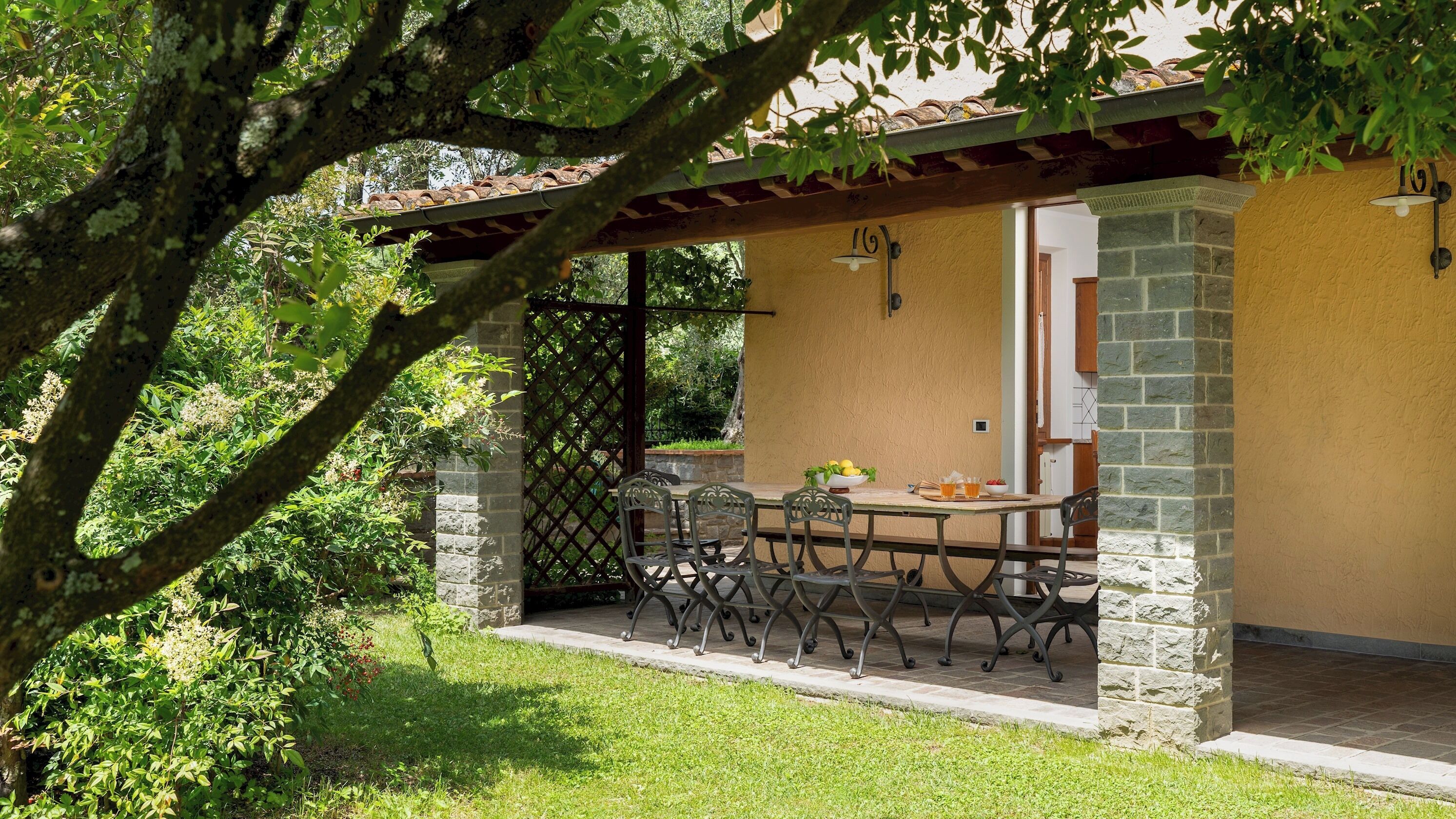 A covered patio with a table and chairs, surrounded by greenery and a stone wall.