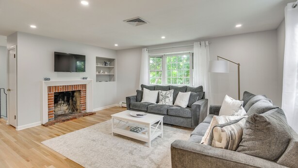 A living room with a fireplace, grey sofas, a white coffee table, and a large window with curtains. - Middletown Vacation Rental: 2 Mi to Beach! (Middletown)